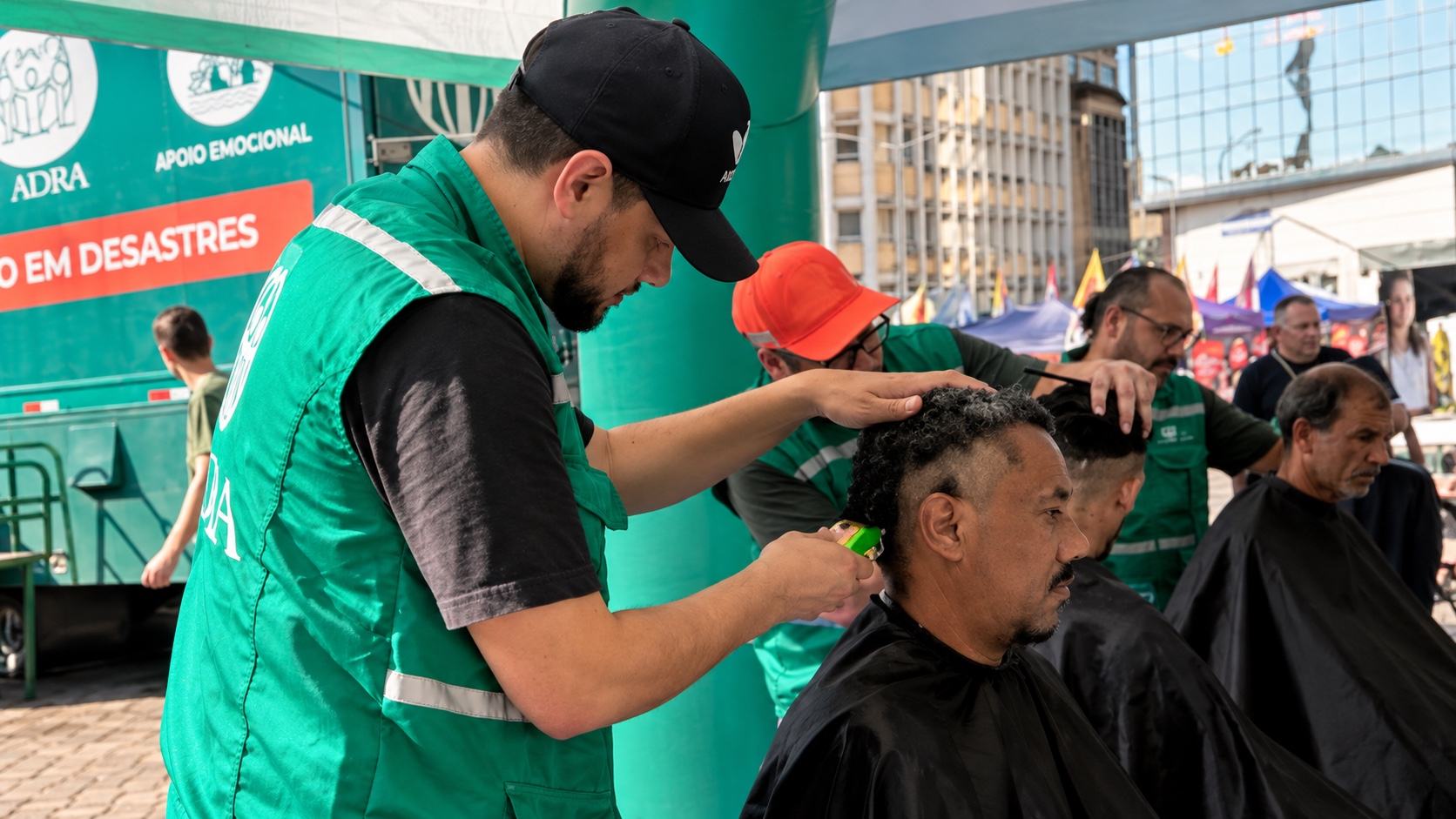Voluntário de colete verde corta o cabelo de um homem sob uma tenda de atendimento na rua