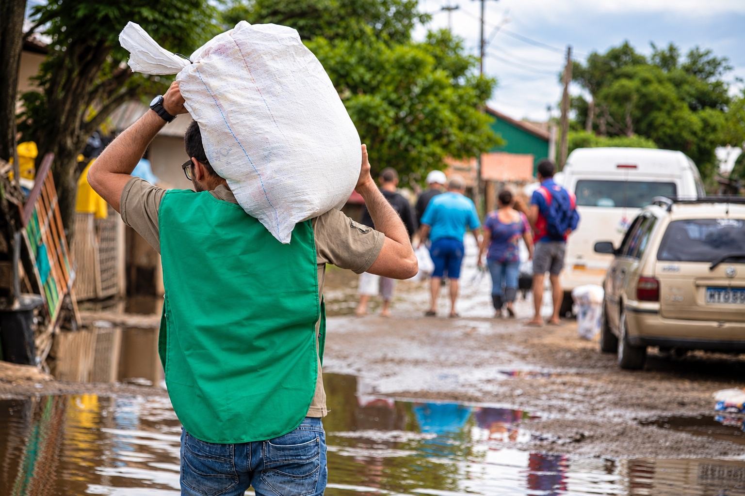 Voluntário de colete verde carrega um saco de doações nos ombros por uma rua alagada